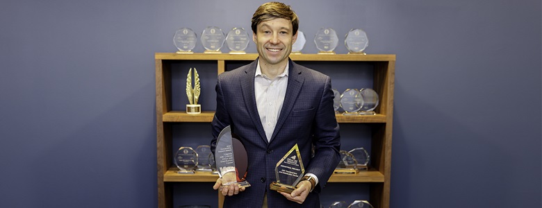 Travis Hill holding two awards with blue background